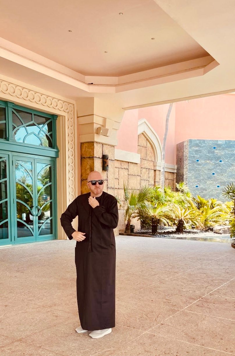 Viktor Grünwald in a dark kandura and sunglasses, standing in front of a hotel entrance in Dubai.